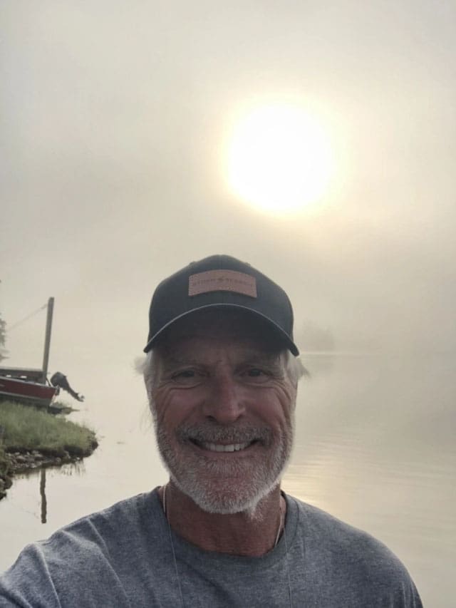 Man in a hat smiles by the water.